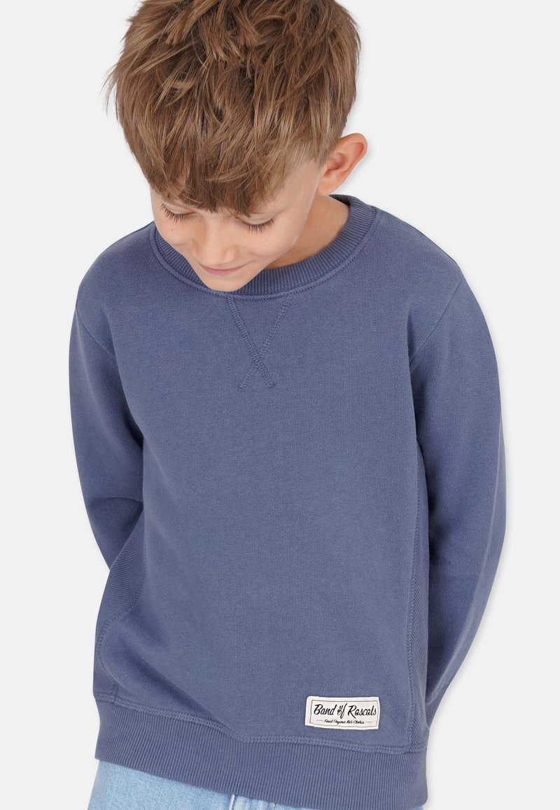 Sininen puuvillapaita, jossa on pyöreä kaula-aukko, resorit hihoissa ja helmassa. Vasemmassa alareunassa on pieni brändilogo.