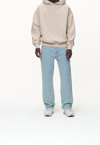 Sweat à capuche beige avec une poche frontale et des lettres, associé à un jean droit bleu clair et des baskets grises. Tenue simple et décontractée.