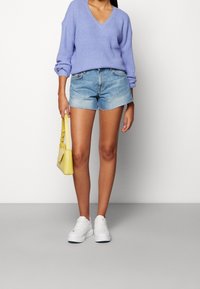 Jasnofioletowy dziergany sweter z dekoltem w serek, niebieskie dżinsowe szorty, białe trampki i mała żółta torebka na tle jednolitego tła.