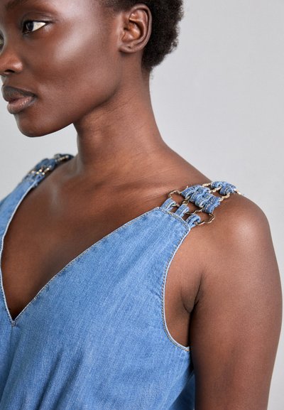 Denimmekko, jossa on syvä V-pääntie, säädettävät punotut hihnat metallikoristeilla ja rento istuvuus. Väri on vaaleansininen, jossa on näkyvät saumat.
