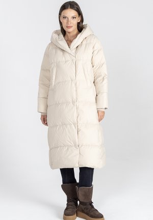 Długi beżowy płaszcz puchowy z kapturem, o pikowanym wzorze, z dwiema zewnętrznymi kieszeniami oraz gładką, błyszczącą powierzchnią. W połączeniu z brązowymi butami.
