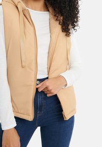 Beige wattierte Weste mit Kapuze und Frontreißverschluss, mit beigen Kordelzügen, getragen über einem weißen Langarmoberteil und dunkelblauen Jeans.