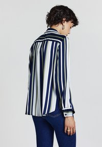 Gestreifte Bluse in Marineblau, Grün und Weiß; hat lange Ärmel, einen Kragen und einen abgerundeten Saum. Der Stoff wirkt glatt mit einem leichten Glanz.