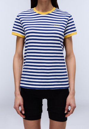 Woman wearing a blue and white striped t-shirt with yellow trim on collar and sleeves, paired with black shorts.