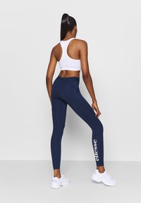 Leggings sportivi blu navy con una superficie liscia, vita alta e un logo bianco sulla coscia sinistra. Abbinati a un reggiseno sportivo bianco con schiena a fascia.