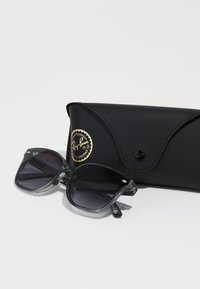 Ochelari de soare Ray-Ban cu un cadru gri transparent și lentile întunecate, însoțiți de un toc texturat negru cu un logo auriu.