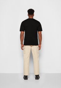 T-shirt noire à manches courtes vue de dos, associée à un pantalon cargo beige et des chaussures de sport noires, sur un fond blanc uni.