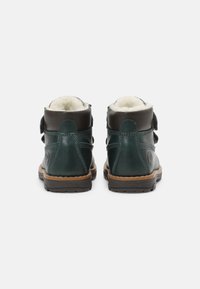 Botas de tobillo de cuero verde con acentos marrones, forro blanco suave y correas de velcro, con una suela marrón texturizada y detalles de costura.