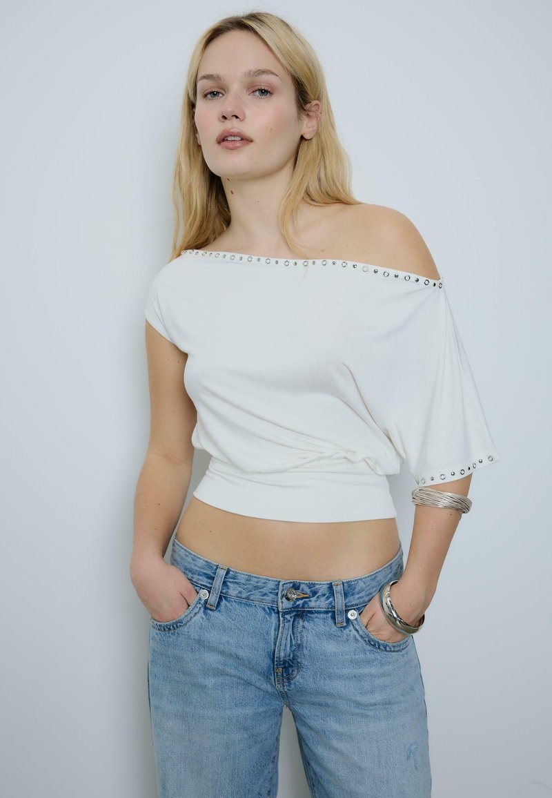 Młoda kobieta w białej bluzce z odkrytymi ramionami z srebrnymi ćwiekami i niebieskich dżinsach, z rękami w kieszeniach, stoi na jasnym, jednolitym tle.