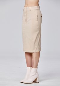 Jupe crayon beige au design épuré, dotée d'une taille avec passants de ceinture, d'une fermeture éclair et de poches latérales. Associée à des bottines blanches. 