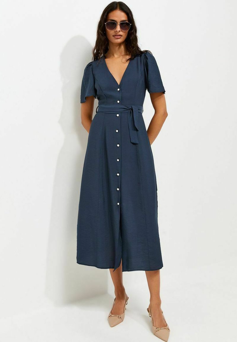 Woman wearing a navy blue button-down midi dress with puff sleeves and a waist tie, beige heels, and dark sunglasses against a plain background.