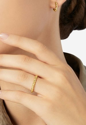 Gold texturierter Creolen-Ohrring und passender Ring, getragen an der linken Hand nahe dem Gesicht, mit Haaren, die zu einem tiefen Dutt gestylt sind, und sichtbarer neutraler Kleidung.