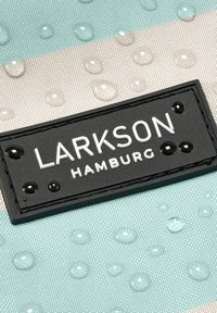 Svart rektangulär lapp med vit text "LARKSON HAMBURG" på en randig tygbakgrund med vattendroppar.