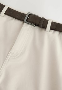 Beżowe spodnie z detalem przeszyć z przodu, zabezpieczone ciemnobrązowym skórzanym paskiem i srebrną klamrą.