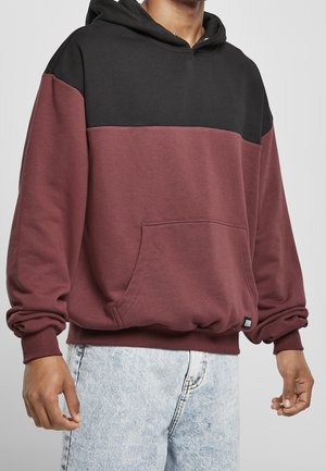 Persoon die een tweekleurige hoodie draagt met een zwart bovenstuk en een bordeauxrode onderkant, gecombineerd met lichtblauwe jeans met een zuuraswassing, staand tegen een effen achtergrond.