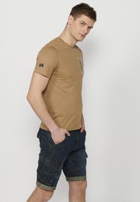T-shirt marron à manches courtes en coton lisse, avec un imprimé graphique sur le devant, associé à un short cargo en denim bleu avec des ourlets roulés.