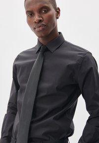 Camisa negra de vestir hecha de tela suave con cuello puntiagudo y puños de botón, combinada con una corbata gris oscuro, que muestra un aspecto formal.
