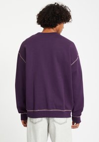 Paarse sweatshirt met een relaxte pasvorm, met contrasterende stiksels en geribbelde manchetten. Het materiaal lijkt zacht met een gladde textuur.