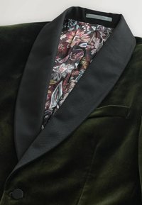 Zielona aksamitna marynarka z czarnym satynowym patką. Wnętrze ma podszewkę w kwiatowy wzór w kolorach burgundowym, czarnym i pastelowych.