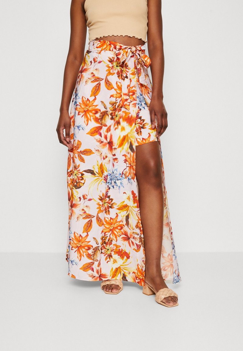 Femme portant un crop top beige et une jupe maxi fendue haute avec des motifs floraux orange, jaune et bleu, associée à des sandales à talons beiges.