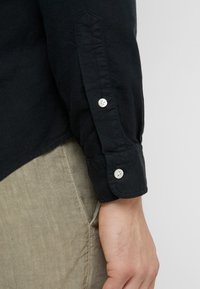 Černá košile na knoflíky s dlouhými rukávy, s bílými knoflíky na manžetách a hladkou bavlněnou texturou. V kombinaci se světle béžovými kalhotami.