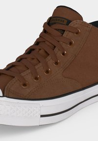 Καφέ ψηλό sneaker Converse με καφέ κορδόνια, λευκή λαστιχένια σόλα και μαύρη γραμμή λεπτομέρειας.