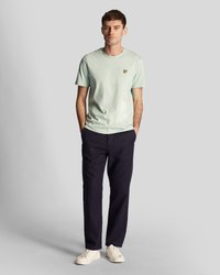 Un homme se tient avec les mains dans les poches, portant un t-shirt vert clair, un pantalon sombre et des sneakers blanches sur un fond gris uni.