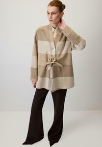 Kardigans ar svītrām bēša un gaiši brūnā krāsā, ar jostu viduklī, nolaistām pleciem un trim pogām, kopā ar tumši brūnām zvana formas biksēm.