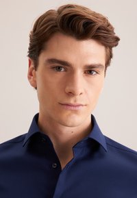 Marineblauwe shirt met een gestructureerde stof, een button-down kraag en subtiele stikdetails, gedragen door een jonge man met kort haar.