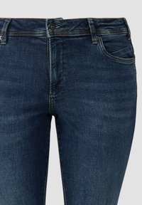 Ciemnoniebieskie dżinsy z denimu mają dopasowany krój, pięciokieszeniowy fason, widoczne przeszycia oraz zapięcie na guzik w talii.