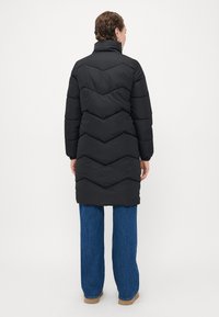 Cappotto puffa nero con colletto alto, caratterizzato da un design a trapunta a zigzag, che arriva a metà coscia, abbinato a jeans a gamba larga blu.