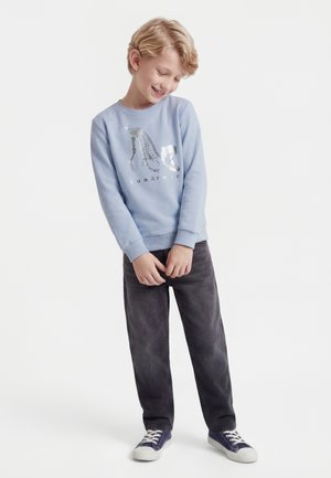 Smilende blond gutt iført en blå genser med dinosaurskjelettprint, mørke bukser og marineblå joggesko, stående mot en hvit bakgrunn.