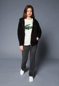 Čierna mikina na zips s zelenými akcentmi, biele tričko s grafikou zeleného aligátora, šedé džínsy, biele tenisky, stojí na šedom pozadí.