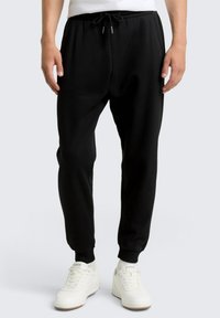 Pantalones de chándal negros con cinturilla elástica y cordón ajustable. Tobillos ajustados y una textura suave. Combinados con zapatillas blancas.