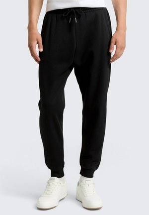 Pantalons de jogging noirs avec taille élastique et cordon de serrage ajustable. Chevilles resserrées et texture douce. Portés avec des baskets blanches.