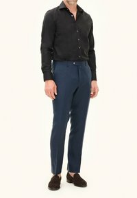 Zwarte overhemd met knopen, blauwe op maat gemaakte broek en donkerbruine instapschoenen. De outfit heeft strakke lijnen en een verzorgde uitstraling.