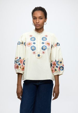 Femme portant un chemisier crème avec une broderie florale bleue, rose et jaune sur la poitrine et des manches bouffantes, associé à un jean bleu foncé.