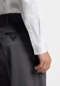 Weißes Hemd mit Knopfmanschetten, kombiniert mit dunkelgrauer, strukturierter Hose mit Karomuster und einer mit Knöpfen versehenen Taschenapplikation.