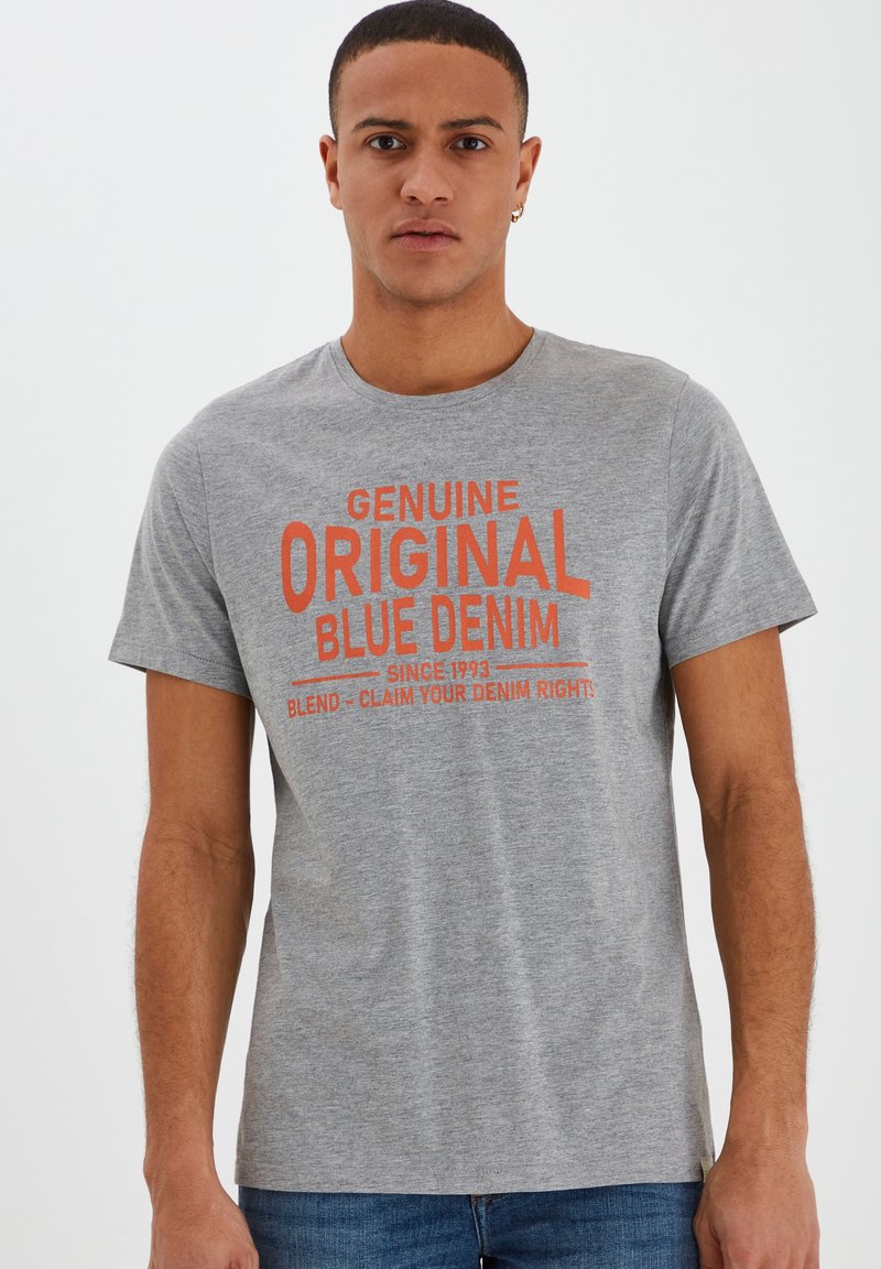 Camiseta de algodón gris con mangas cortas, que presenta un texto naranja en negrita: "DENIM AZUL ORIGINAL GENUINO" y detalles adicionales de marca.