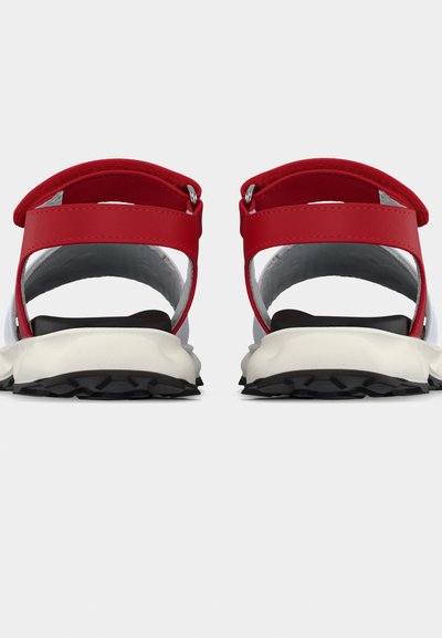 Ζευγάρι κόκκινες και λευκές σανδάλες με ρυθμιζόμενα λουριά στον αστράγαλο, μαύρες σόλες με υφή, φαίνονται από πίσω σε απλό φόντο.