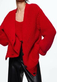 Punainen neulepusero, jossa on löysä istuvuus, syvä V-pääntie ja tekstuuri. Ominaisuuksina suuret mustat napit ja kerrostettu muotoilu yhteensopivan paidan päällä.