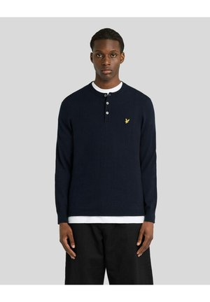 Ung mand iført en navyblå langærmet henley-shirt med tre knapper og et lille gult fuglelogo på brystet.