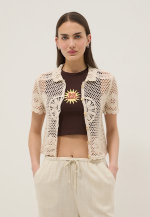 Donna che indossa una maglietta beige a maniche corte all'uncinetto sopra un crop top marrone con grafica del sole "Sol Mate" e pantaloni beige chiaro con coulisse.