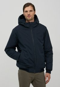 Giacca isolante blu navy con zip frontale, cappuccio e polsini elastici. Presenta una texture liscia e tasche laterali per il contenimento.