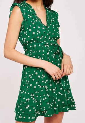 Frau trägt ein ärmelloses grünes Kleid mit weißem Gänseblümchenmuster, gerafftem Ausschnitt, Knöpfen und einem gebundenen Taillengürtel.