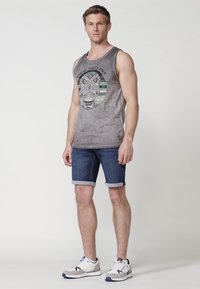 Siva majica brez rokavov z grafiko lobanje, v kombinaciji s temno modrimi denim kratkimi hlačami z zavihanimi robovi in belimi športnimi copati s sivimi detajli.