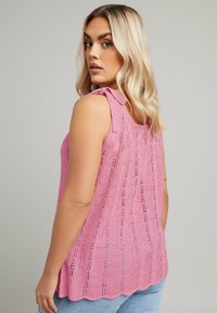 Rosa ärmlös topp med en texturerad, öppen stickad mönster och rosett-detaler på axlarna, med en scalloped kant.