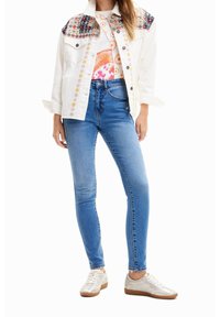 Bílá džínová bunda s vzorovanými rameny, spárovaná s modrými skinny džíny a světlými teniskami. Bunda má květinovou výšivku a detaily s knoflíky.