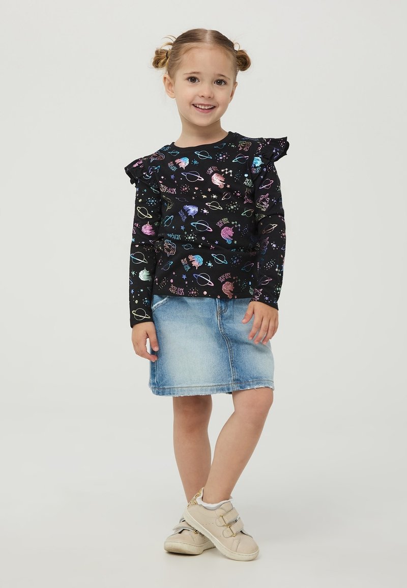 Czarna koszula z długim rękawem z kolorowymi nadrukami w motywy kosmiczne i falbaniastymi akcentami, w połączeniu z jasnoniebieską dżinsową spódnicą i beżowymi trampkami.