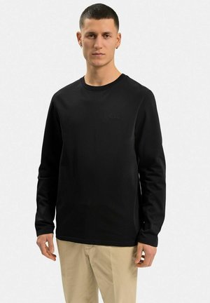 Sort langærmet t-shirt lavet af bomuld, med rund halsudskæring og et subtilt logo på brystet, parret med lyse bukser.